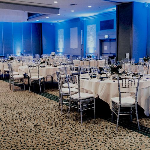 A banquet hall with round tables set for an event, featuring white tablecloths, silver chairs, and blue ambient lighting, creating an elegant atmosphere.