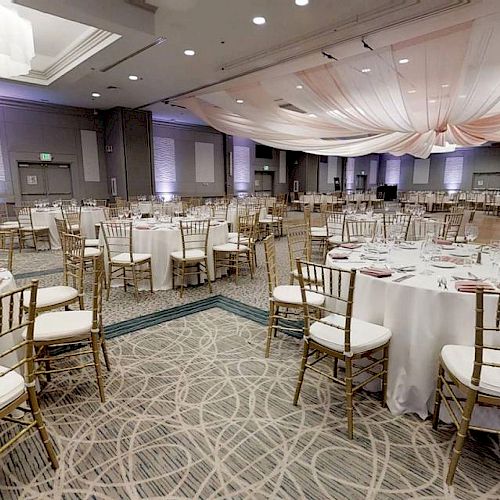 An elegant banquet hall is set up with round tables, gold chairs, white tablecloths, and draped ceiling decorations, prepared for an event.