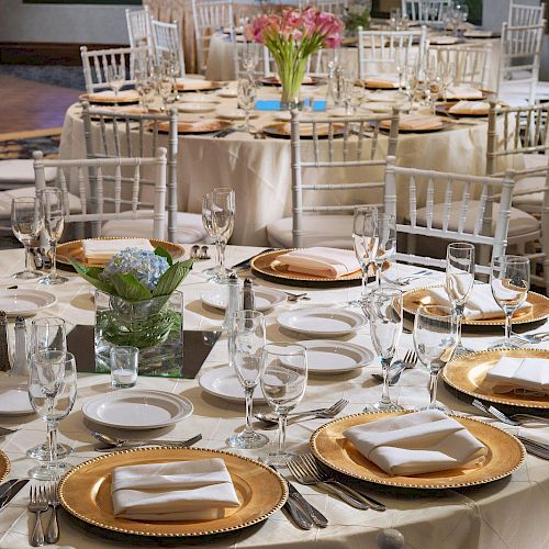 The image shows an elegant banquet setup with round tables adorned with white tablecloths, gold charger plates, neatly folded napkins, and floral centerpieces.
