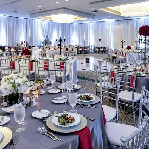 This image shows an elegant banquet hall set up for an event, with round tables, silver chairs, floral centerpieces, and plated salads on each table.