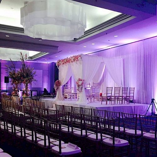 The image shows a beautifully decorated wedding venue with purple lighting, a floral arrangement, and rows of chairs aligned facing a stage with a canopy.