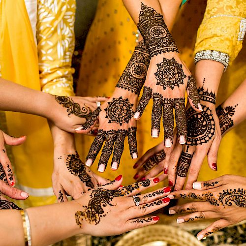 A group of people displaying their beautifully intricate henna designs on their hands, all reaching toward the center in a celebratory gesture.