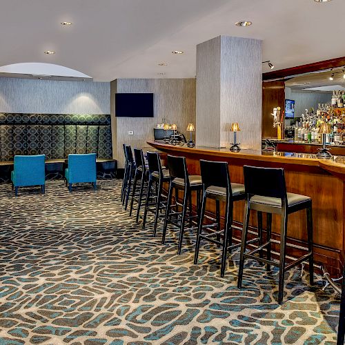 This image shows a modern bar area with a long counter, bar stools, a seating area with blue chairs, and a patterned carpet on the floor.