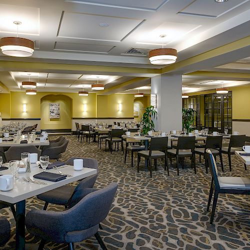 The image shows a modern, well-lit restaurant with neatly arranged tables and chairs, set for dining, featuring a patterned carpet and yellow walls.