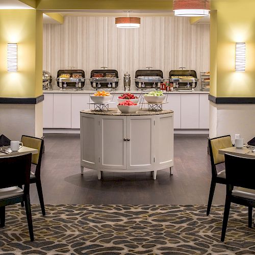 This image shows a restaurant buffet setup with chafing dishes and various food items. Tables are set for dining, under warm lighting.