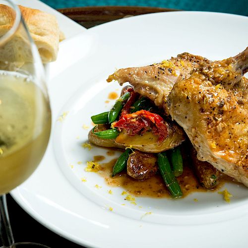 The image shows a plated dish with roasted chicken, green beans, and potatoes, accompanied by a glass of white wine and some bread.