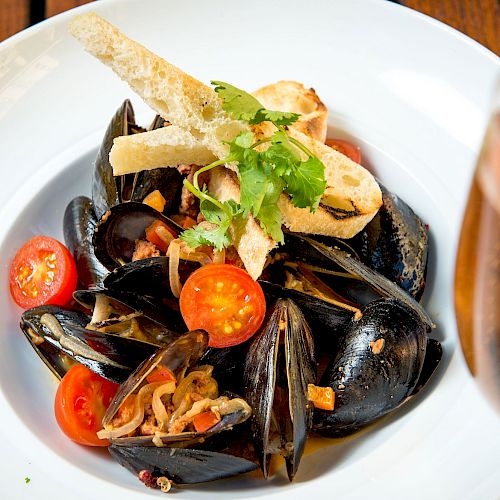 A dish of mussels with sliced cherry tomatoes, garnished with herbs and breadsticks, next to a glass of sparkling beverage, elegantly presented.