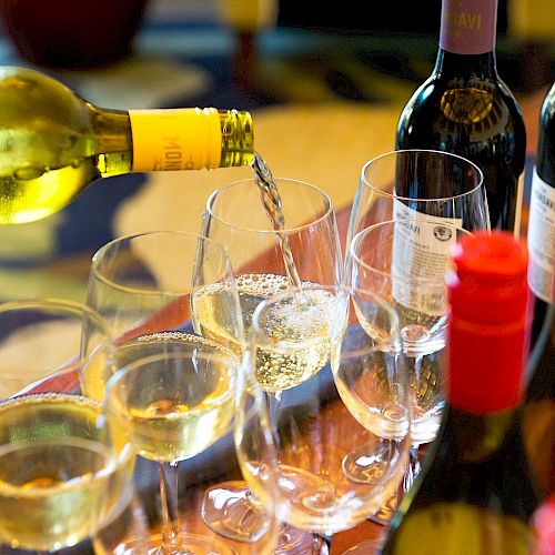 A person is pouring white wine into several wine glasses, with different wine bottles visible on the table.