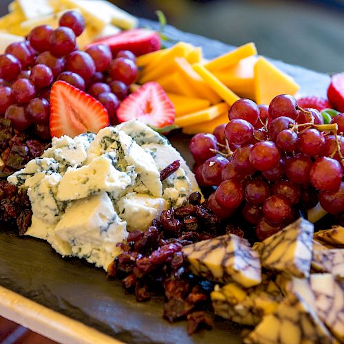 The image shows a charcuterie board with various cheeses, red grapes, strawberries, and dried fruits, arranged on a black slate.