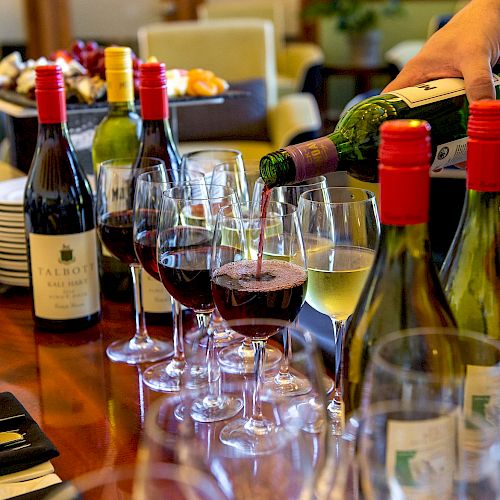 This image shows a person pouring wine into glasses, with several wine bottles and glasses arranged on a wooden table, ready for a gathering.