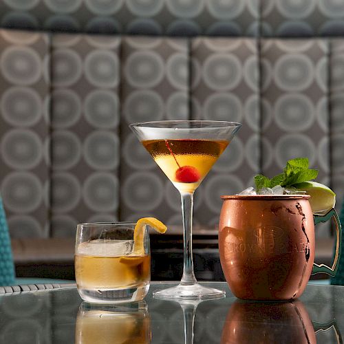 Three drinks on a reflective table: a cocktail with a lemon garnish, a martini with a cherry, and a copper mug with mint and lime.