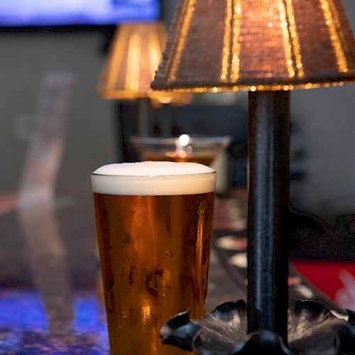 A pint of beer sits on a bar next to a table lamp with a warm glow; a TV screen is visible in the blurred background.