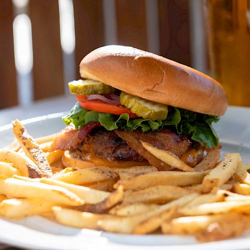 A plate with a burger topped with lettuce, tomato, and pickles, accompanied by fries. A tall glass of beverage is in the background.