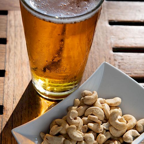 A glass of beer is on a wooden table next to a white dish filled with cashews.