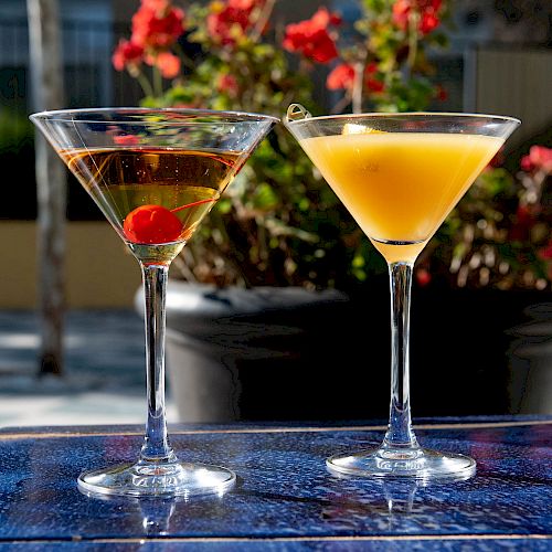 Two martini glasses, one with a cherry and the other with an orange slice, are set on a blue table with a floral background.