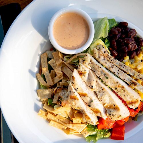 A white bowl contains grilled chicken strips, black beans, corn, tortilla strips, lettuce, red bell pepper, and a cup of pink sauce.