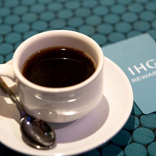 A cup of black coffee sits on a saucer with a spoon, next to an IHG Rewards card on a blue patterned surface.